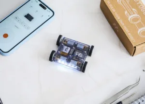 Tiny transparent robotic car with four wheels sits on a white surface next to a blue smartphone displaying controls and a connected app window.