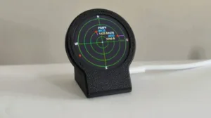 Round radar display on a black stand with green concentric circles and compass directions N, S, E, W showing target data text in the center. Full setup on a table.