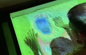 Two children touch a glowing, green-lit sand panel with a blue circular center at an interactive exhibit.