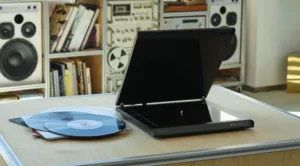 Open turntable with blue vinyl records on a light wood table, retro stereo gear and books on shelves in the background.