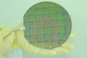 Gloved hands hold a circular silicon wafer with intricate microchip patterns against a light background.