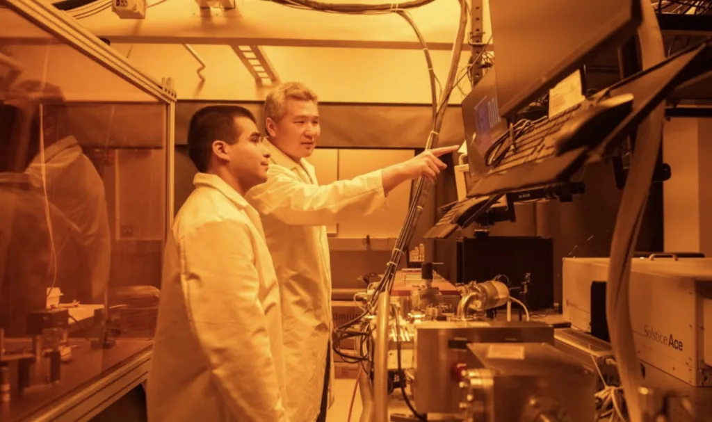 Two technicians in lab coats stand at a workstation, one pointing at a computer screen while operating a large analytical instrument in a bright lab.