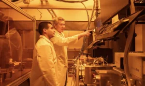 Two technicians in lab coats stand at a workstation, one pointing at a computer screen while operating a large analytical instrument in a bright lab.
