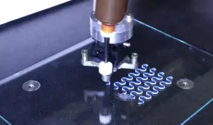 Close-up of a robotic tool forming blue S-shaped springs on a dark worktable.