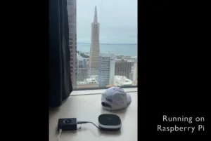 View of a city skyline from a window, featuring a tall triangular skyscraper, with a desk in the foreground and a Raspberry Pi setup nearby.