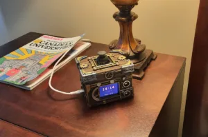 Steampunk-inspired wooden gadget with visible gears and a small blue digital clock showing 14:14 on a brass-wood casing, on a wooden table with a lamp nearby and a colorful magazine beside it.