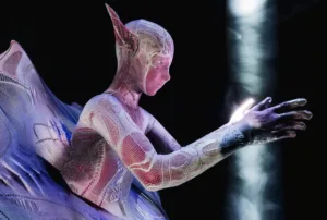 Dancer in a purple lace mesh bodysuit with wing-like fabric, arms extended against a dark background.
