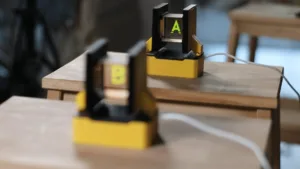 Yellow clamp device with a black frame showing a bright green 'A' on a small screen, placed on a wooden table, blurred foreground in a workshop setting.