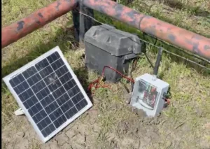 Outdoor off-grid power setup: a solar panel connected to a battery box and a metal control box on muddy ground.