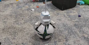 White robotic toy with a soccer-ball patterned body and a vertical antenna standing on a gray speckled carpet.