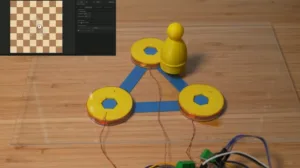 Three yellow spool-like motors wired together to form a triangle on a wooden table, with a yellow peg figure at the top vertex and exposed electronics nearby.