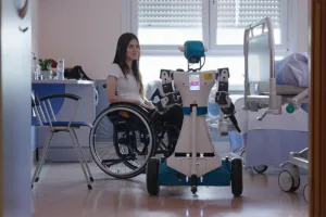Woman in a wheelchair sits in a hospital room beside a blue-and-white service robot with arms folded by its sides.