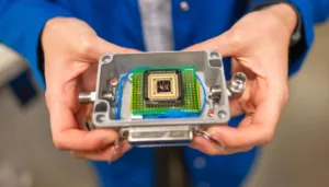Person holds an open metal enclosure showing a green circuit board with a central square IC chip inside a device housing.