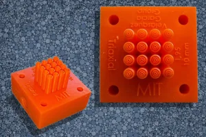 Orange silicone block with a 4x4 grid of small cylindrical pins; shown from a side angle and a top view with 'MIT' molded on the surface.