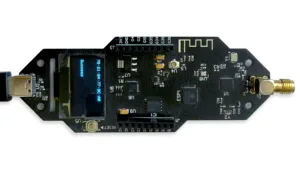 Black IoT development board with a small blue OLED display, USB port on the left, header pins in the center, and a clip-on/wireless antenna on the right.