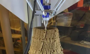 Industrial 3D printer nozzle extruding beige, rope-textured vase-shaped object.