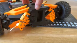 Close-up of an orange robotic chassis; a finger points to a hinge/connector on the black frame near the rear wheel, with a grid ruler in the background.