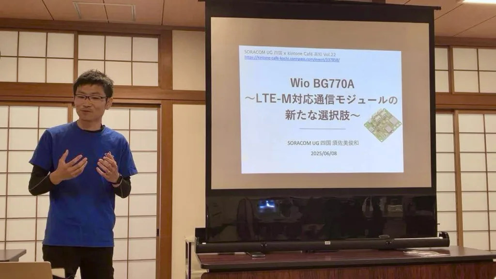 Male presenter in a blue shirt explaining a slide about Wio BG770A LTE-M on a projector screen in a meeting room.
