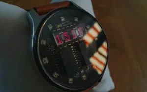 Close-up of a smartwatch internals, showing a circuit board and a pink LED readout reading 16500 labeled 'Battery Indicator'.