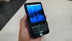 Hand holding a black smartphone with a full QWERTY keyboard and blue wallpaper.