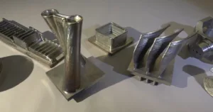 Assorted shiny metal 3D-printed parts laid on a light surface, including a tall curved vase-like piece and stackable square channels.