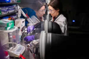 Two researchers wearing safety goggles and purple gloves work at a lab bench inside a fume hood, handling samples.