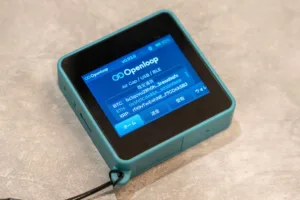 Teal handheld device displaying a blue screen with 'Openloop' app and cryptocurrency wallet options (BTC, ETH, XRP).