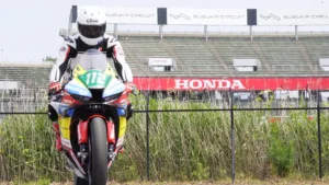 Motorcycle racer on a red-and-blue bike roaring toward the camera on a track with Suzuka Circuit signage in the background and tall grass in the foreground.