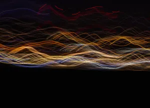 Long-exposure photo of colorful light trails weaving across a dark night sky over a distant horizon, creating abstract waves.