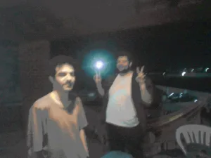 Two men standing at night by the water; one holds a lit phone/flash and the other shows a peace sign, city lights glimmer in the distance.