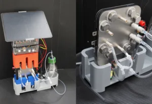 Bench-top lab pump system with tubing, metal manifolds, and an orange heat sink on a gray base. A bottle is connected by clear tubing on the side.