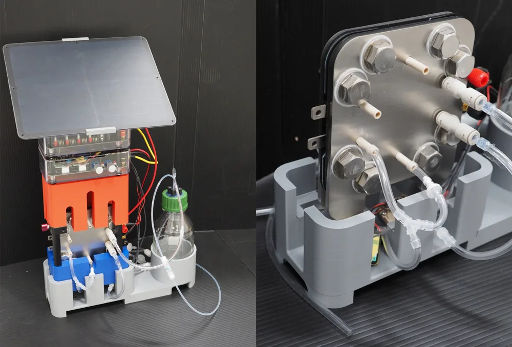 Bench-top lab pump system with tubing, metal manifolds, and an orange heat sink on a gray base. A bottle is connected by clear tubing on the side.