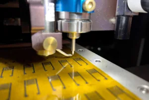 CNC milling machine cutting a yellow circuit-board-like material with a spinning spindle and a steel guide, on a grid-marked worktable