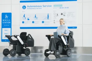 Woman in a motorized wheelchair at a WHILL station beside a blue signage board with 'Autonomous Service' information in a bright lobby.