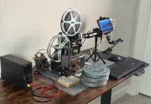 Desk workspace with a vintage film projector on a stand, two reels, wired equipment, and a monitor on a tripod nearby.